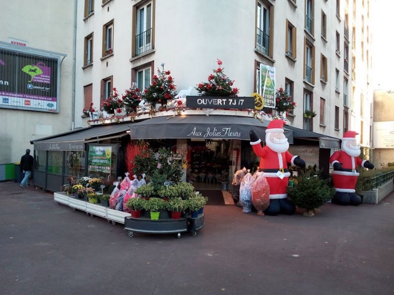 La devanture d’Aux Jolies Fleurs décorée pour Noël&nbsp;: store gris, «&nbsp;Ouvert 7j/7&nbsp;», sapins et plantes sur le trottoir